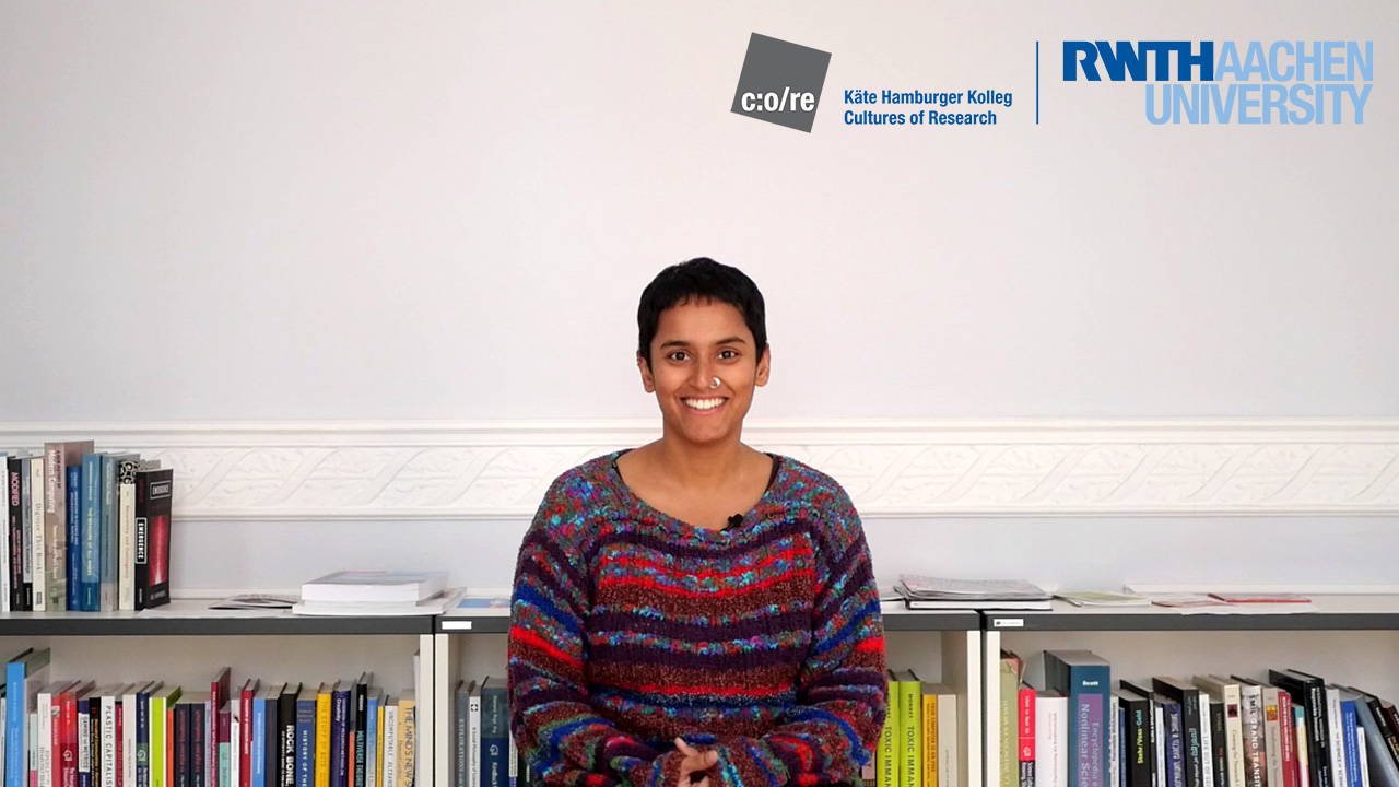 Woman wearing multicolor sweater and glasses, smiling in front of a bookshelf.