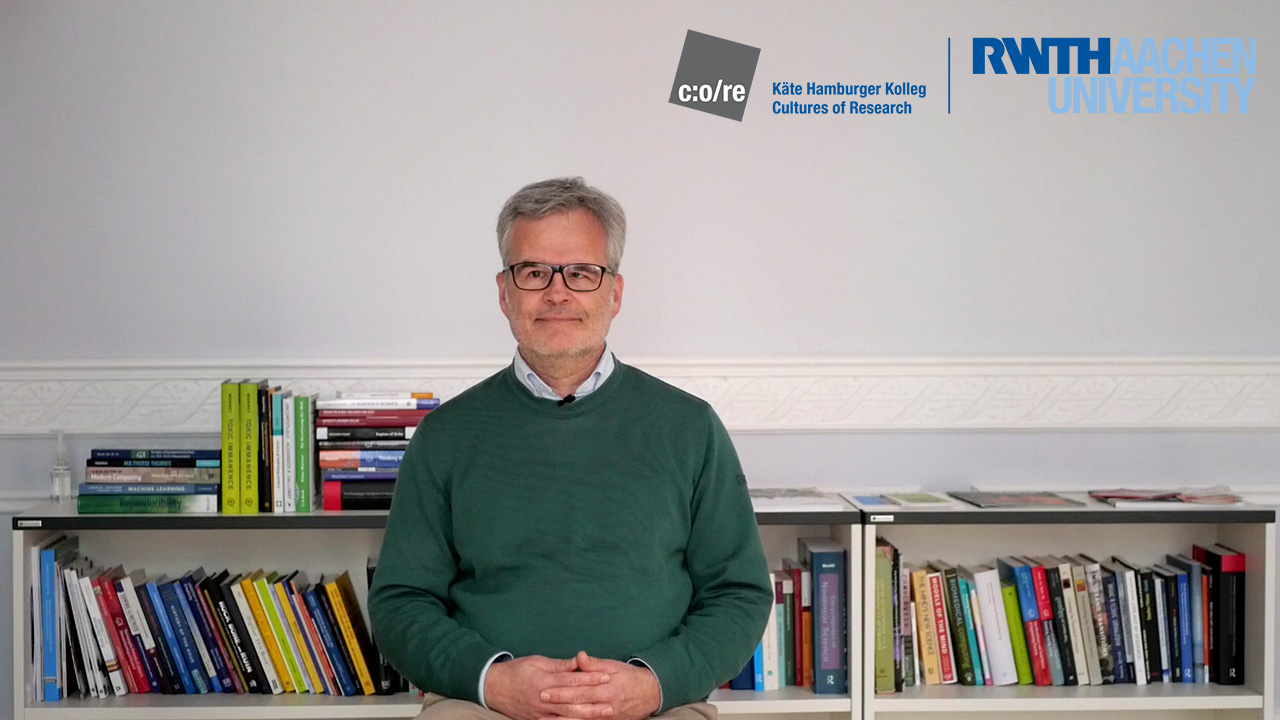 Man wearing green sweater and glasses, sitting in front of a bookshelf.