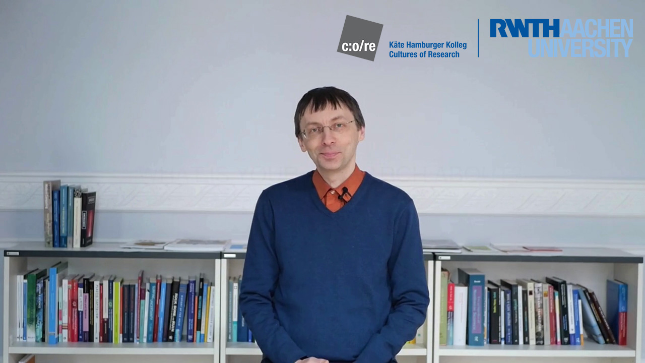 Man wearing blue sweater and glasses, sitting in front of a bookshelf.