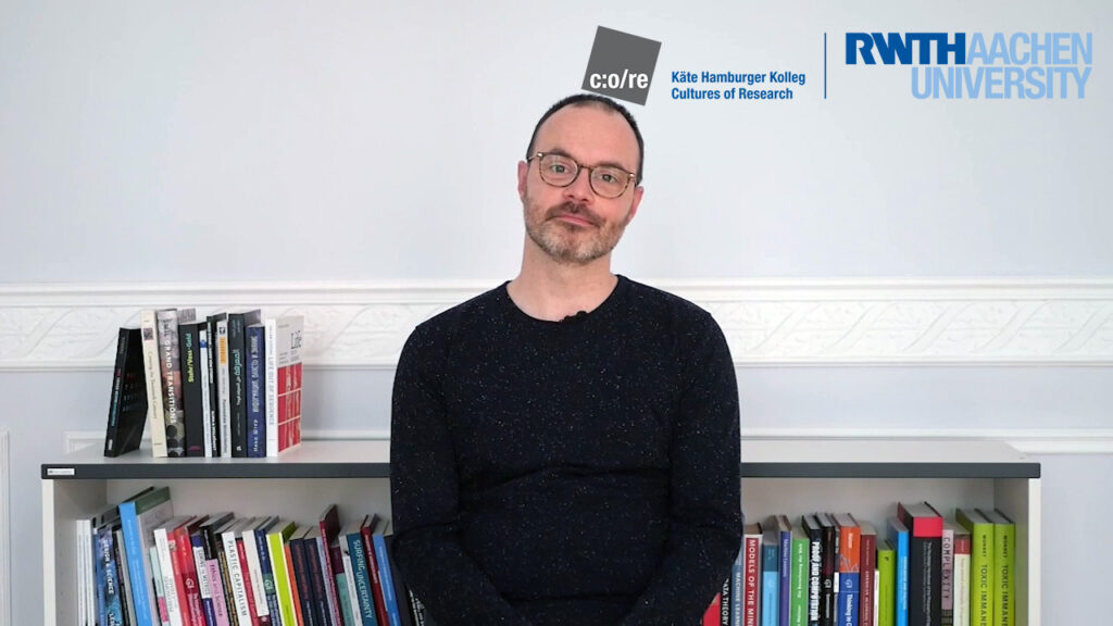 Man wearing black sweater and glasses, sitting in front of a bookshelf.