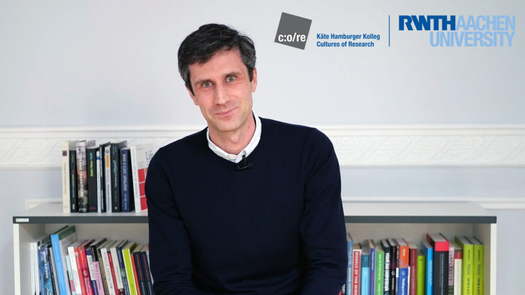 Man wearing black sweater, sitting in front of a bookshelf.