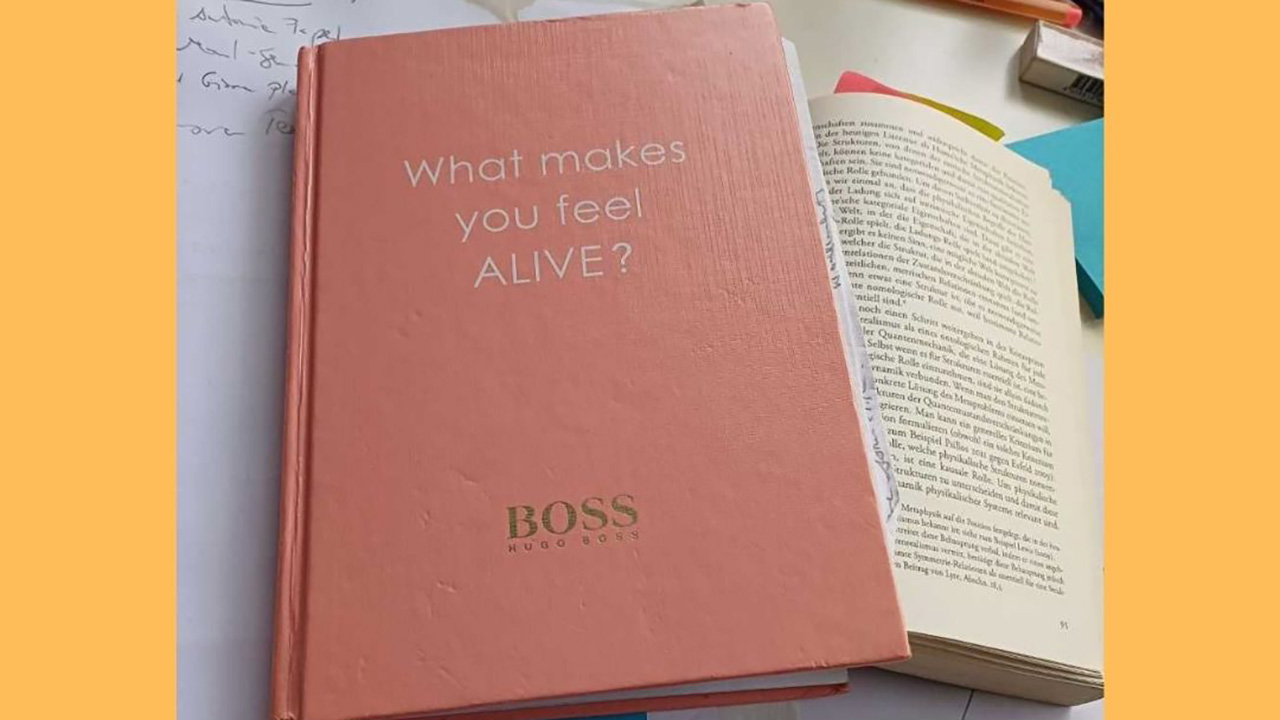 A pink notebook with the words "What makes you feel alive?" written on it, on an open book and several papers with handwritten notes.