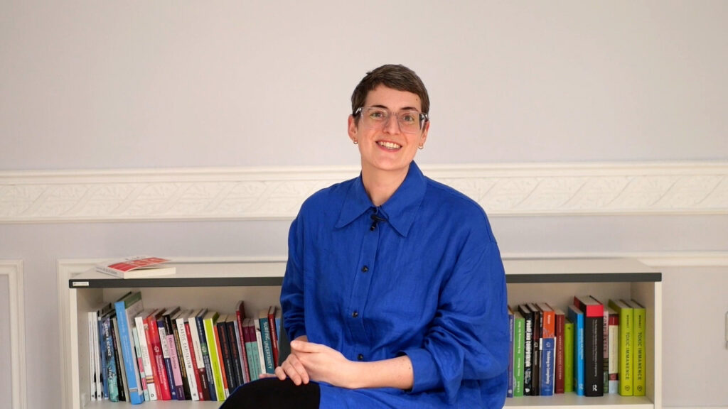 Smiling woman wearing blue shirt and glasses, sitting in front of a bookshelf.