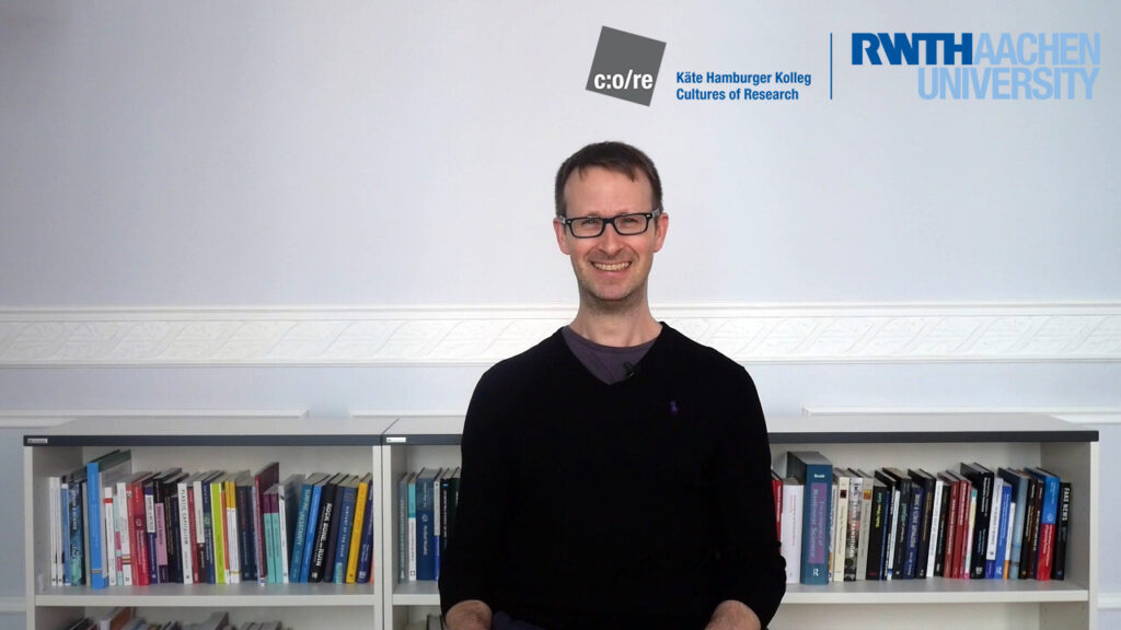 Man wearing black sweater and glasses, sitting in front of a bookshelf.