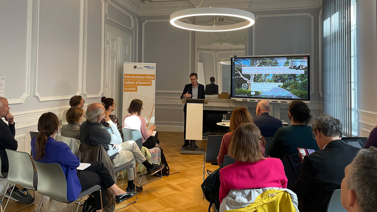Man wearing suit presenting at a podium, beside a large screen, in front of an audience seated in rows of chairs in a small, ornate lecture hall.