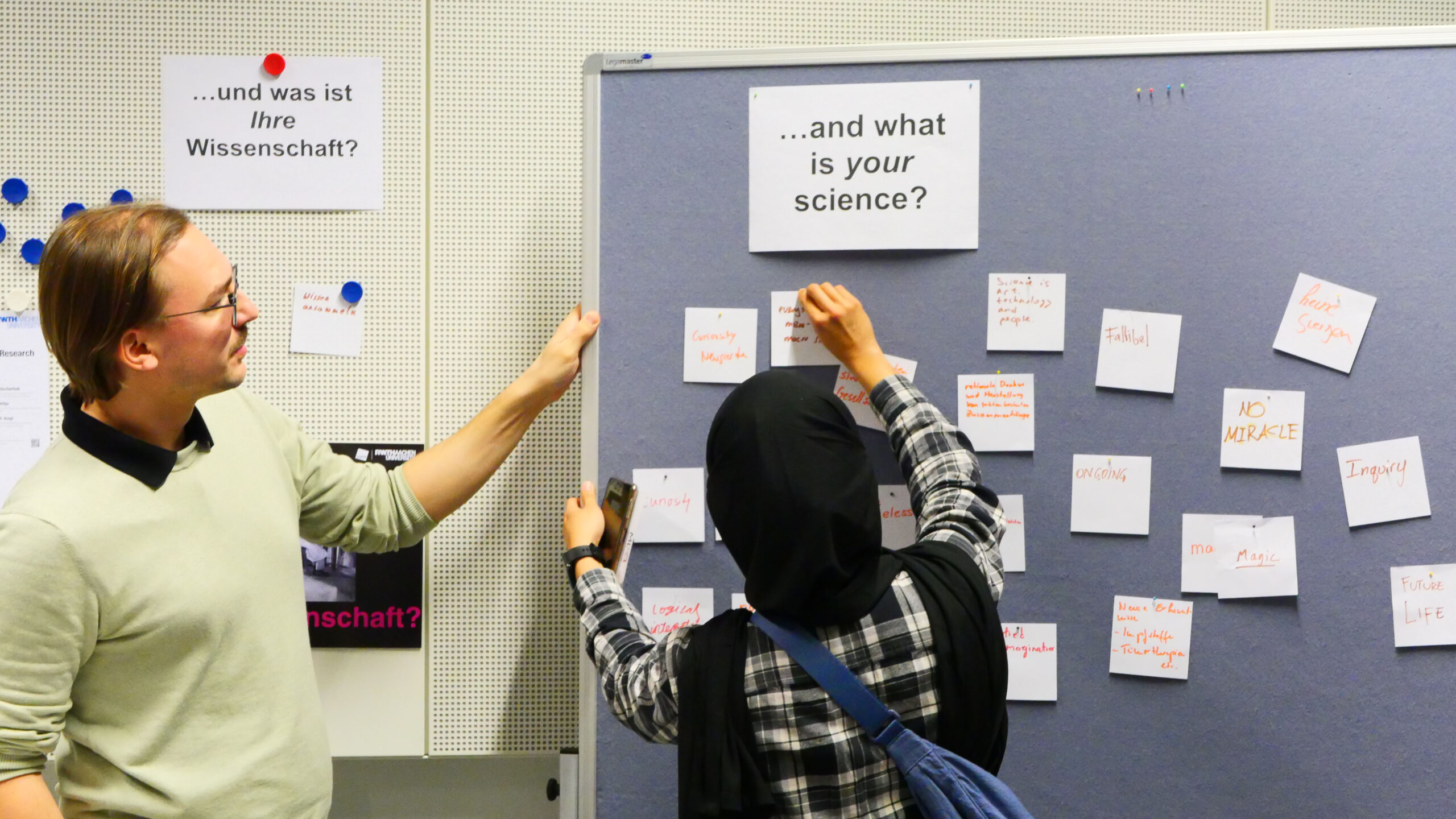 Participant pinning handwritten note to a large bulletin board with the question "...and what is your science?" on it, next to a man holding the board.