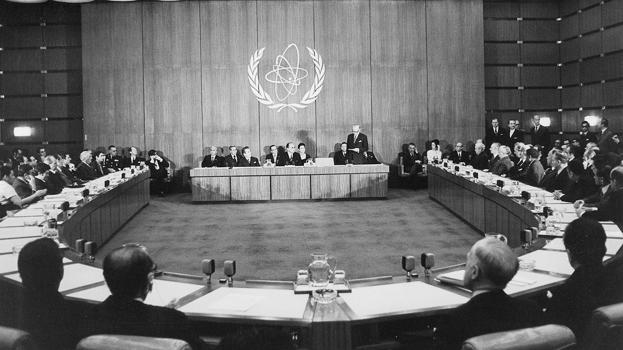 Black and white photograph of an international diplomatic assembly held in a large conference hall, delegates seated around a U-shaped table facing a central podium, the emblem of IAEA displayed on the wall.