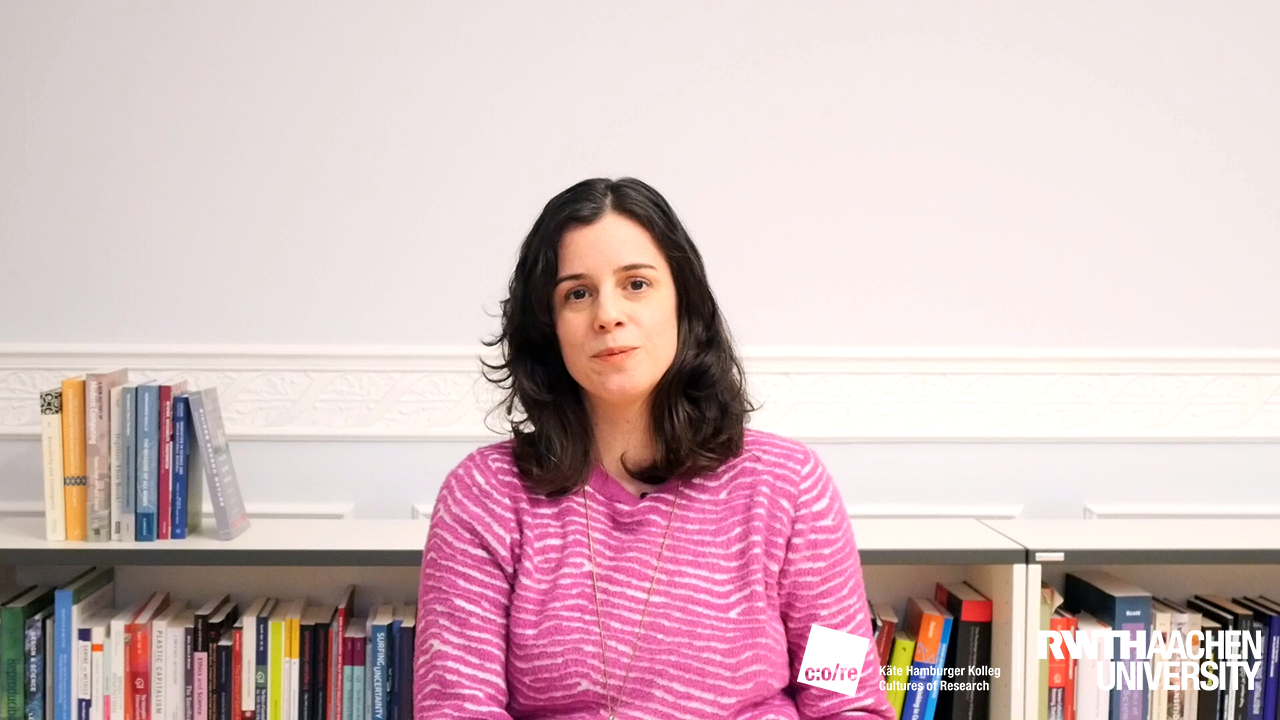 A female person waering a pink blouse, sitting in front of a bookshelf.