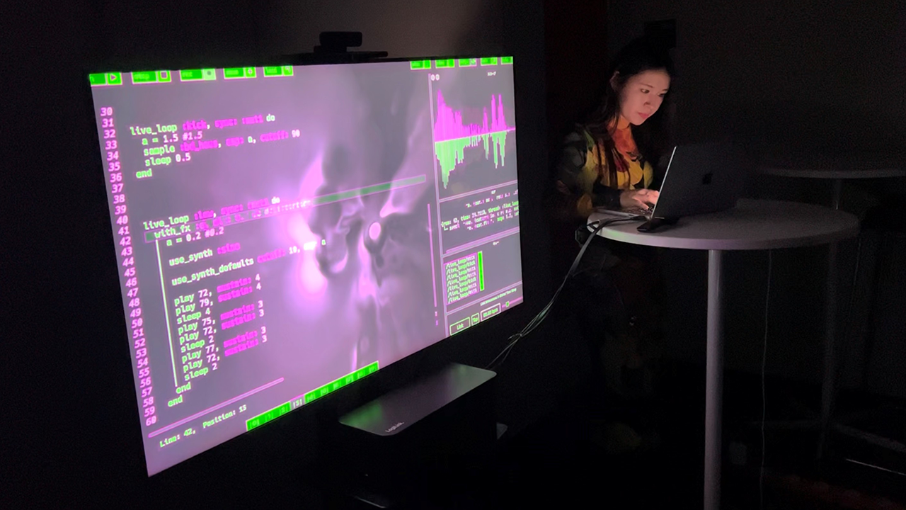 A female person presenting behind a computer, beside a large purple-lit monitor.