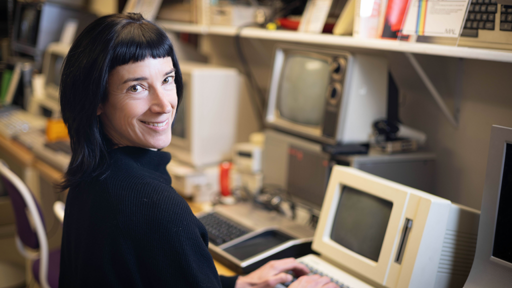 Woman in black at a vintage computer station with CRT monitors, smiling at camera.
