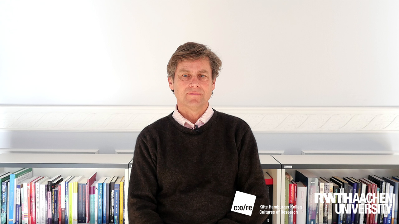 Man in black sweater, posing in front of bookshelf.