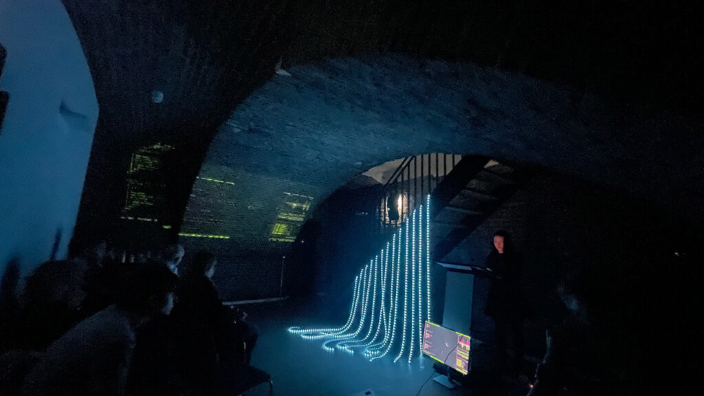 Dark exhibition space under an arched brick ceiling, featuring an art installation with a monitor and strands of blue light tubes cascading down from a staircase.