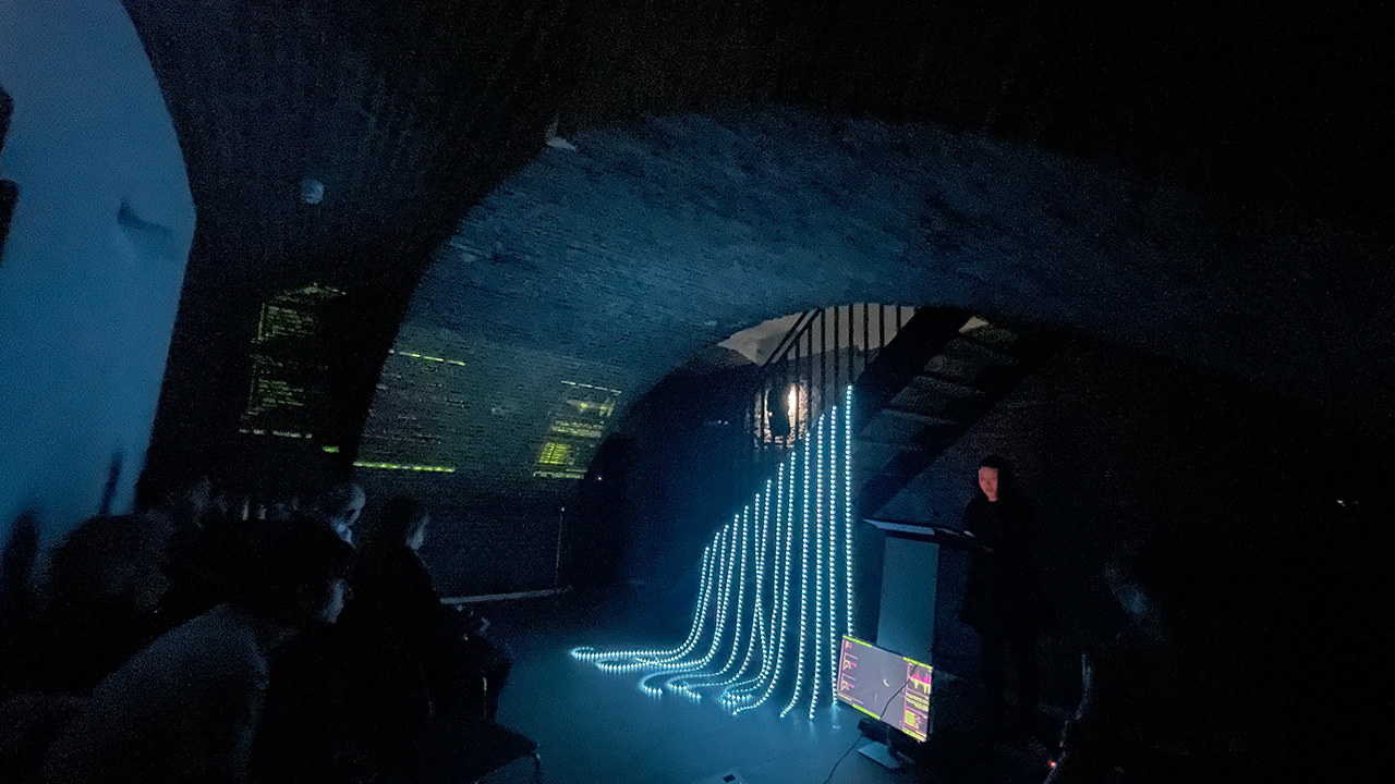 Dark exhibition space under an arched brick ceiling, featuring an art installation with a monitor and strands of blue light tubes cascading down from a staircase.