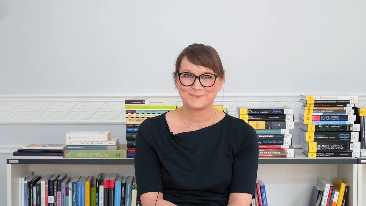 Portrait of a woman with black glasses and in a black shirt sitting in front of a bookshelf.