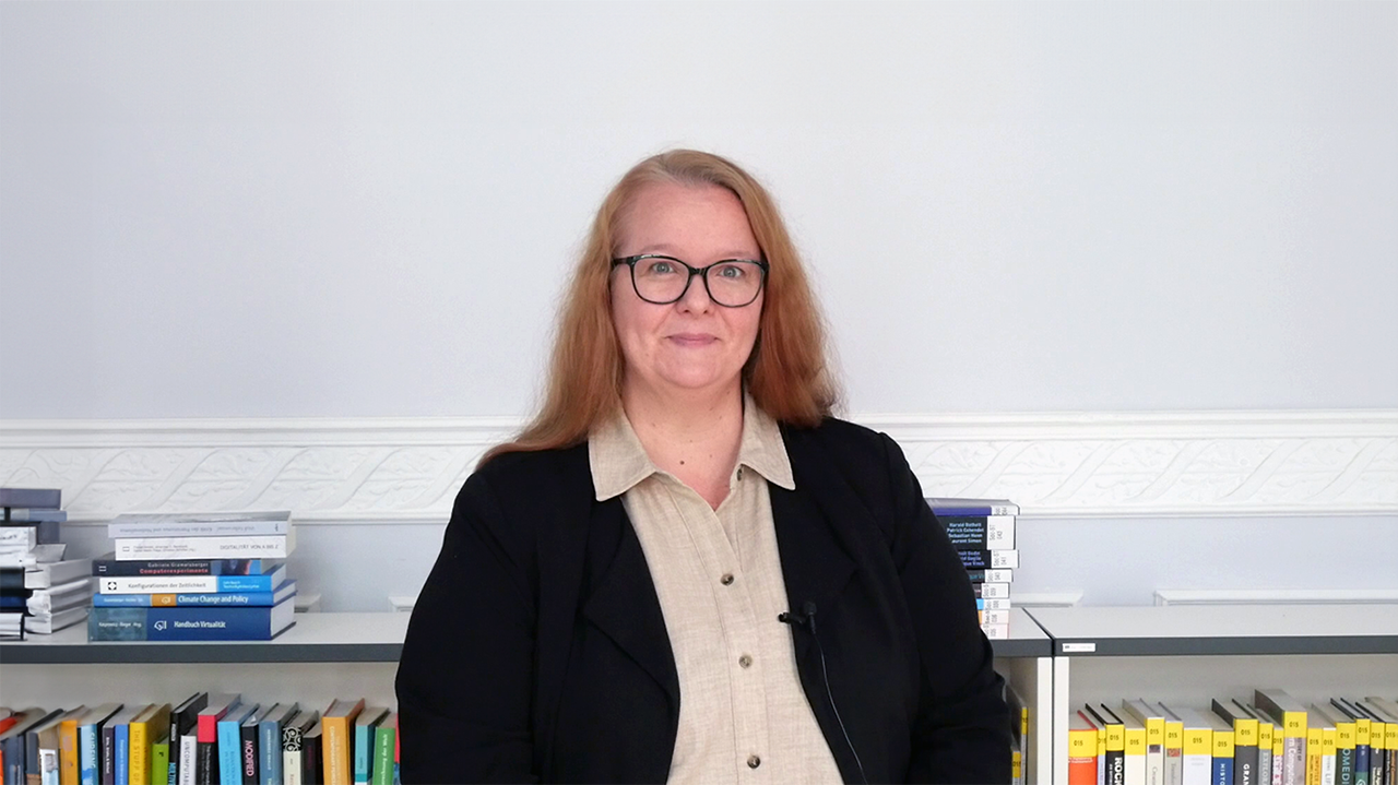 Portrait of a woman with black glasses, a beige blouse and a black cardigan sitting in front of a bookshelf.