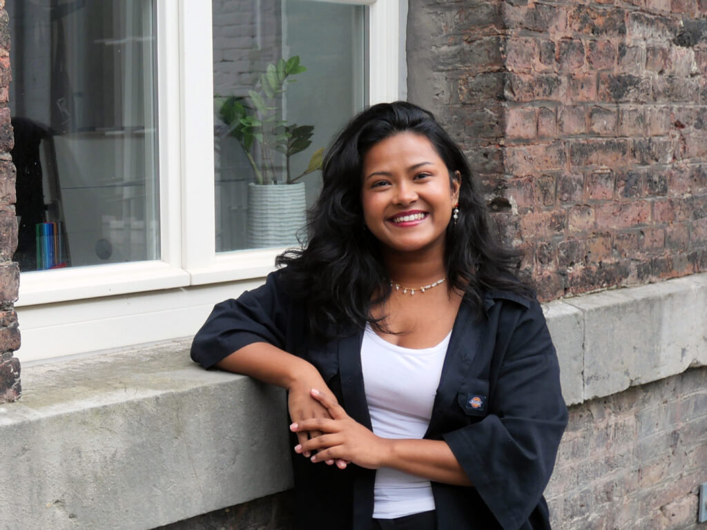 A woman standing in front of a brick wall and smiling at the camera. Seh is waering a black jacket over a white top and accerssorized with earrind and a necklace